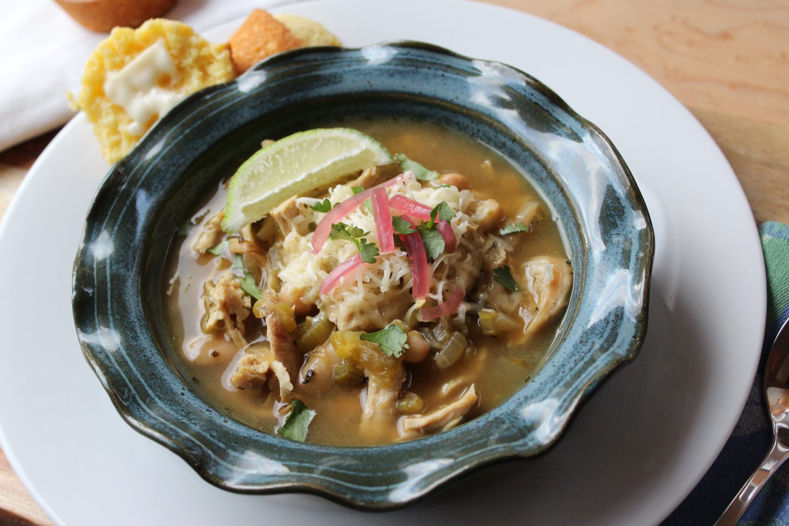 Horizontal photo of my original recipe for White Bean Chicken Chili. Blue pottery bowl with ruffled edge filled with white chicken chili and garnished with cilantro, lime and cilantro. The bowl is placed on a white plate with a buttered corn muffin with a spoon and white napkin to the side.