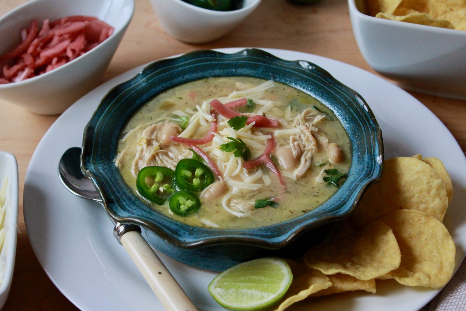 Horizontal photo of Our Favorite White Chicken Chili updated. Blue pottery bowl with ruffled edge filled with white chicken chili and garnished with cilantro, sliced fresh jalapeños and cilantro. The bowl is surrounded by limes, jalapeños, picked red onions, tortilla chips and it placed on a white plate with a spoon and a tan colored napkin.