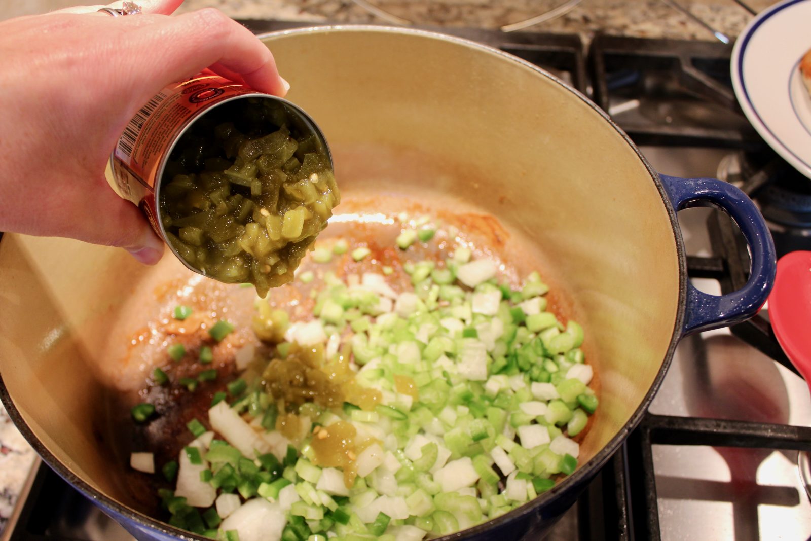 Recipe photo for White Chicken Chili showing adding a can of chopped green chilies to a blue LeCreuset dutch oven with chopped celery, onion and jalapeño already in pot.  Pot is on a gas stove.
