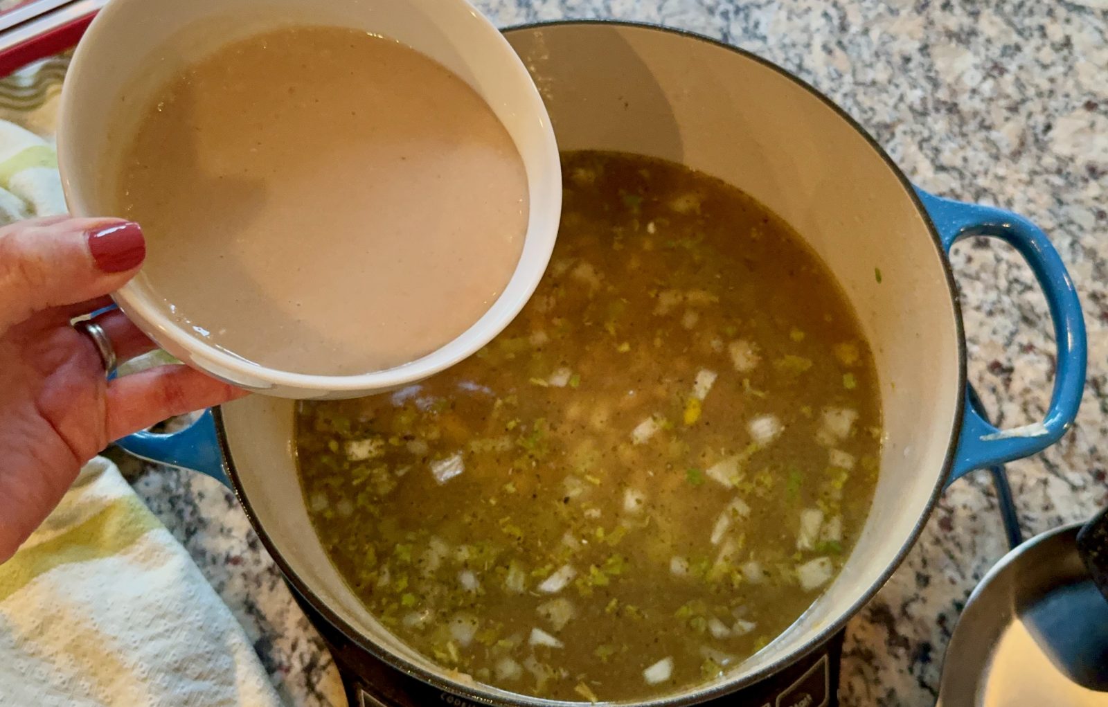 Recipe photo for White Chicken Chili showing adding a white bowlful of pureed cannellini beans into a blue Le Creuset dutch oven with the simmering broth and vegetables in the pot. The pot is on a gas stovetop.