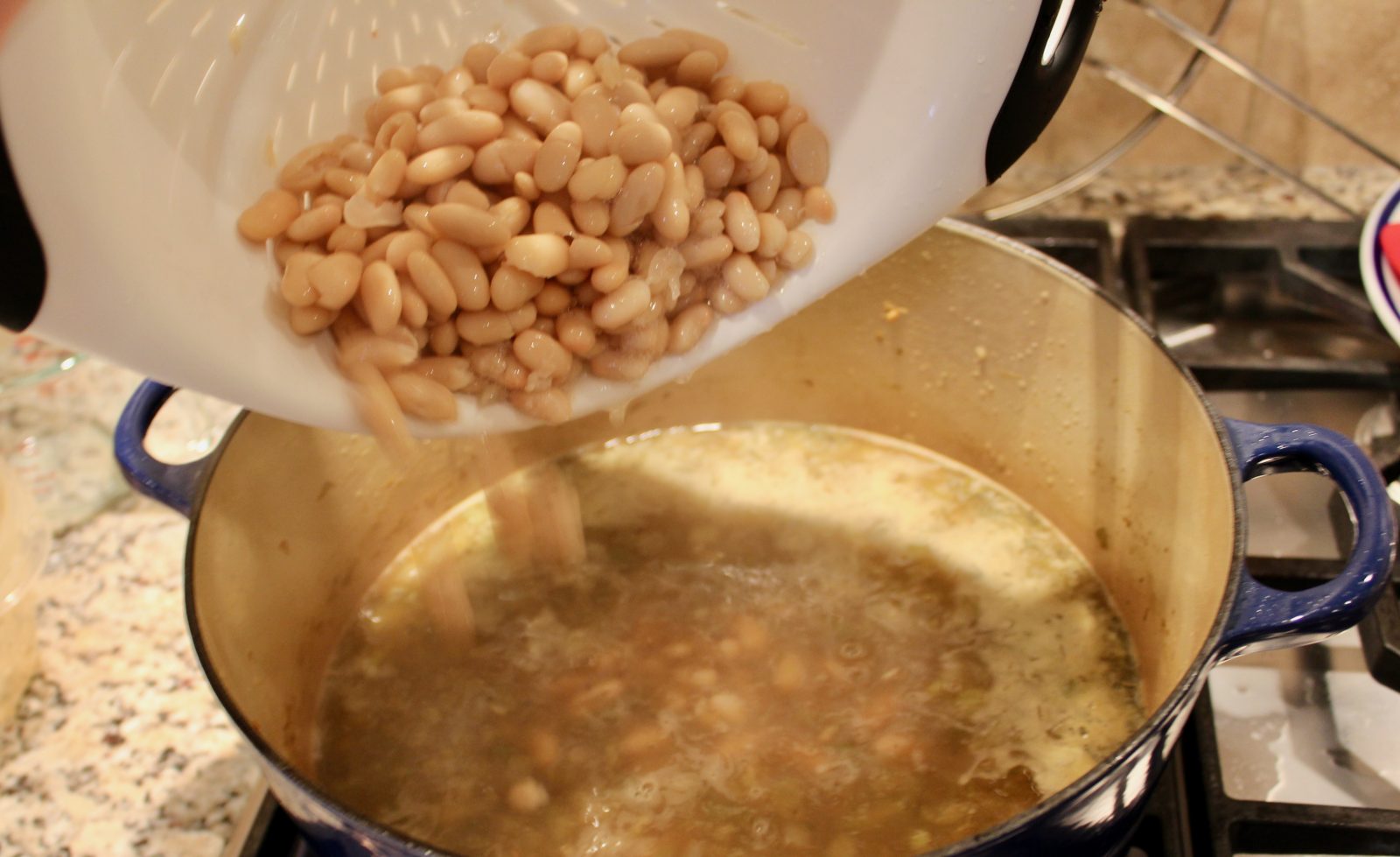 Recipe photo for White Chicken Chili showing adding cannellini beans from a white collander into a blue Le Creuset dutch oven with the simmering broth and vegetables in the pot. The pot is on a gas stovetop.