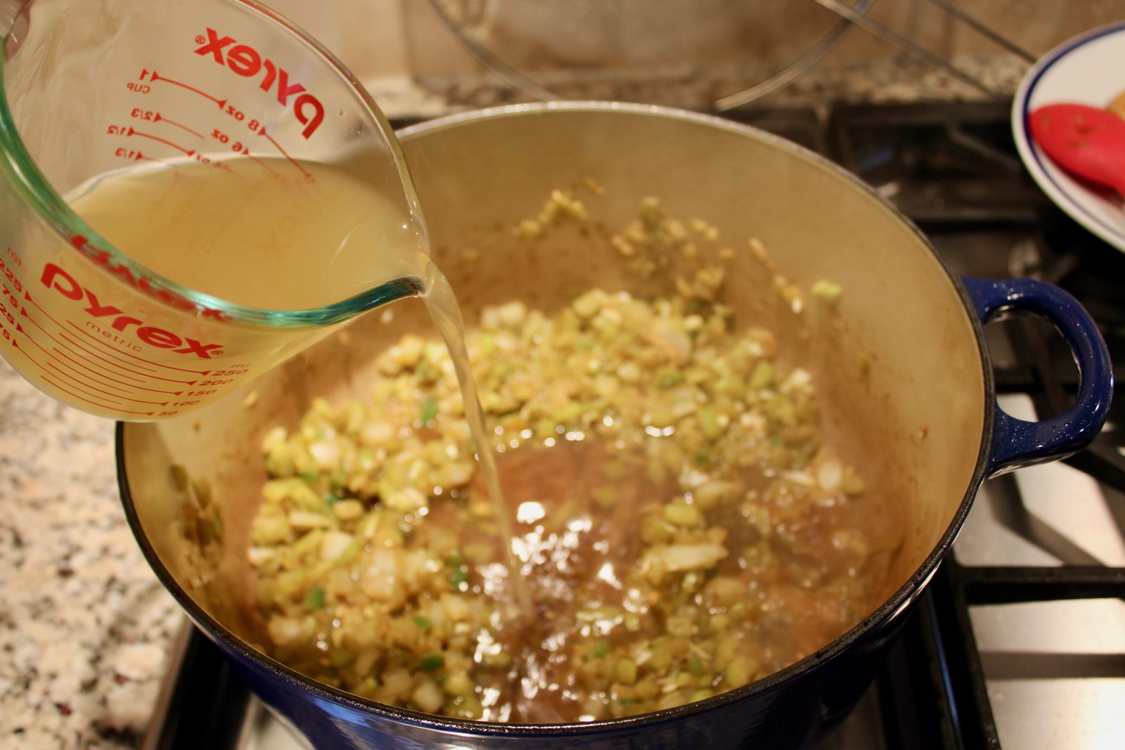 Recipe photo for White Chicken Chili showing adding chicken broth from a glass pyrex measuring cup into a blue Le Creuset dutch oven with sautéed onion, celery, jalapeños and green chilies in the pot.  The pot is on a gas stovetop.
