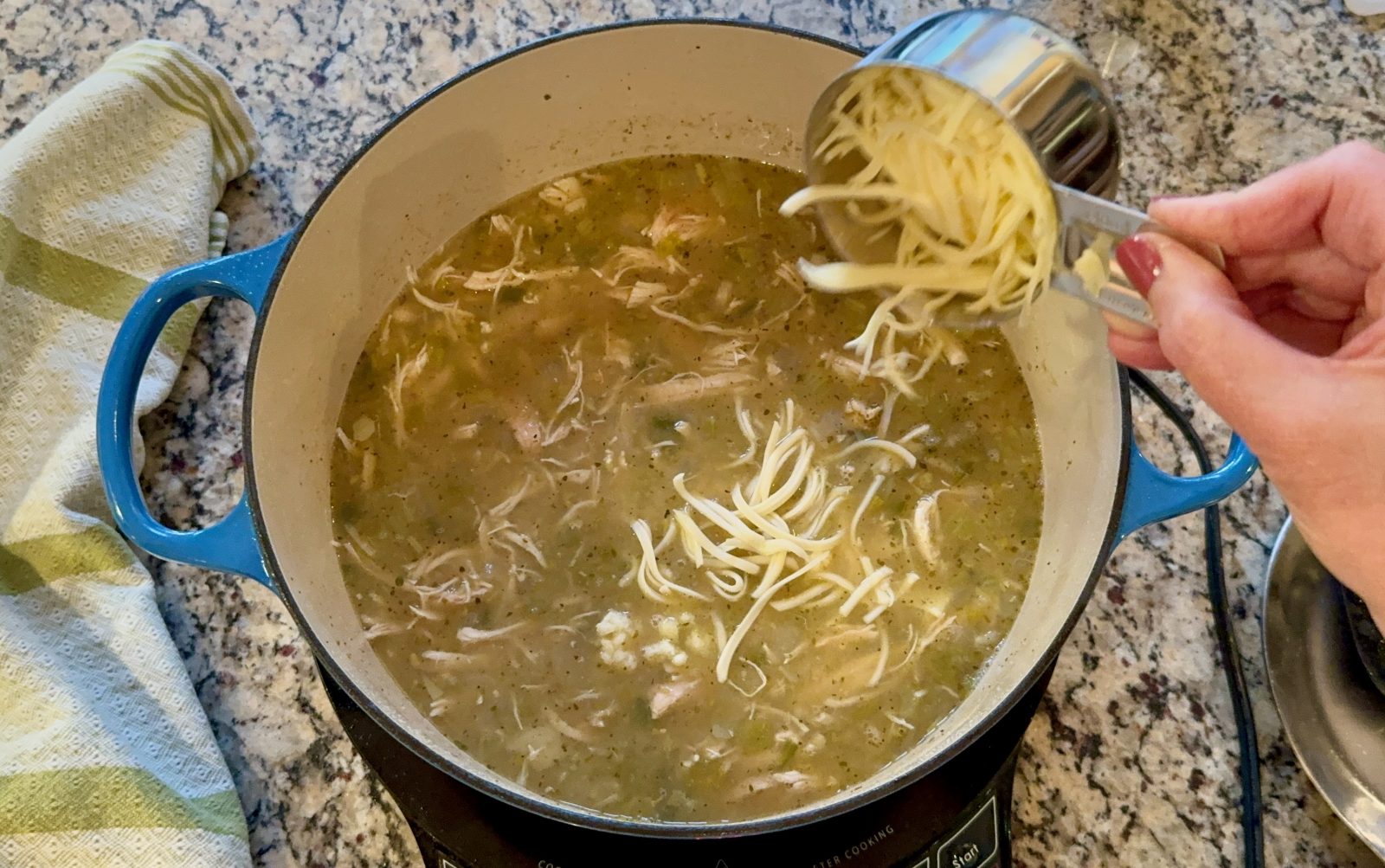Recipe Photo of White chicken chili.  Blue Le Creuset pot with simmering white chicken chili in it.  Adding shredded Monterey jack cheese from 1 cup stainless steel measuring cup.  