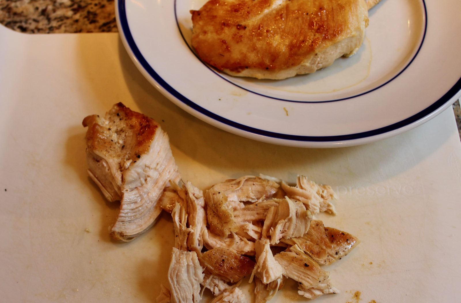 Recipe photo for White Chicken Chili showing shredded cooked chicken breast.  One breast is shredded on a white cutting board while the other breasts are whole on a white plate with a blue rim.
