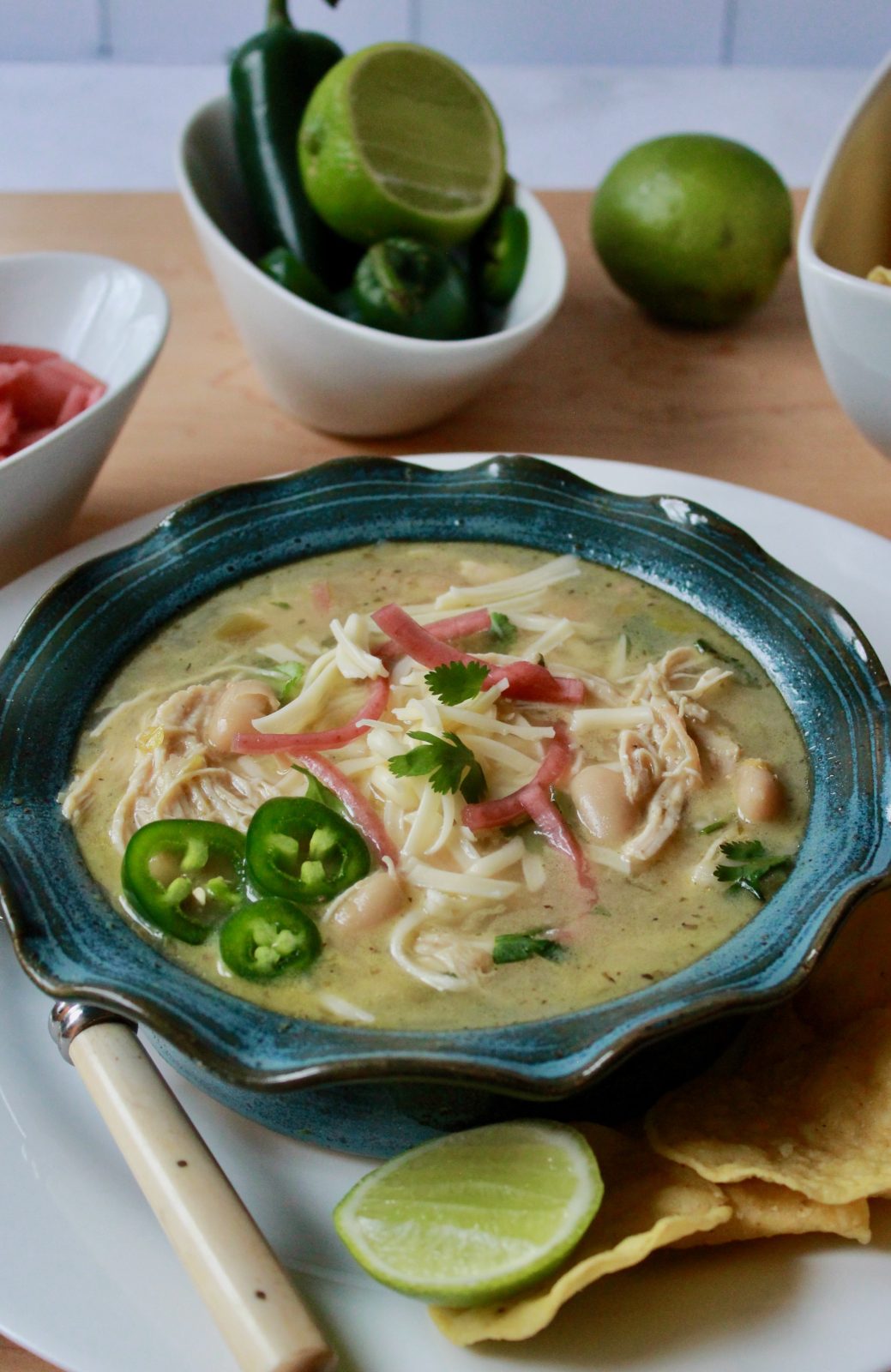 Vertical photo of Our Favorite White Chicken Chili updated. Blue pottery bowl with ruffled edge filled with white chicken chili and garnished with cilantro, sliced fresh jalapeños and cilantro. The bowl is surrounded by limes, jalapeños, picked red onions, tortilla chips and it placed on a white plate with a spoon.
