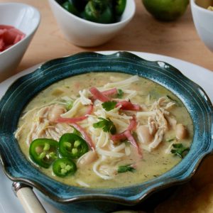Vertical photo of Our Favorite White Chicken Chili updated. Blue pottery bowl with ruffled edge filled with white chicken chili and garnished with cilantro, sliced fresh jalapeños and cilantro. The bowl is surrounded by limes, jalapeños, picked red onions, tortilla chips and it placed on a white plate with a spoon.