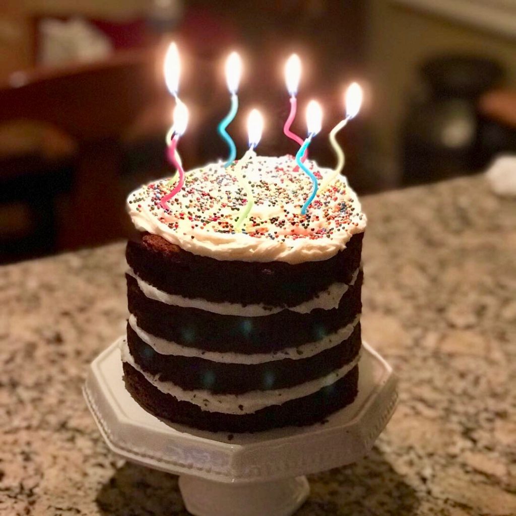Melted Ice Cream Cake on white pedestal with lit birthday candles, 2 6-inch layers cut in half and filled with homemade vanilla buttercream. Cake has naked sides.  Cake made with Devil's Food Cake Mix and Melted Chocolate Ice Cream