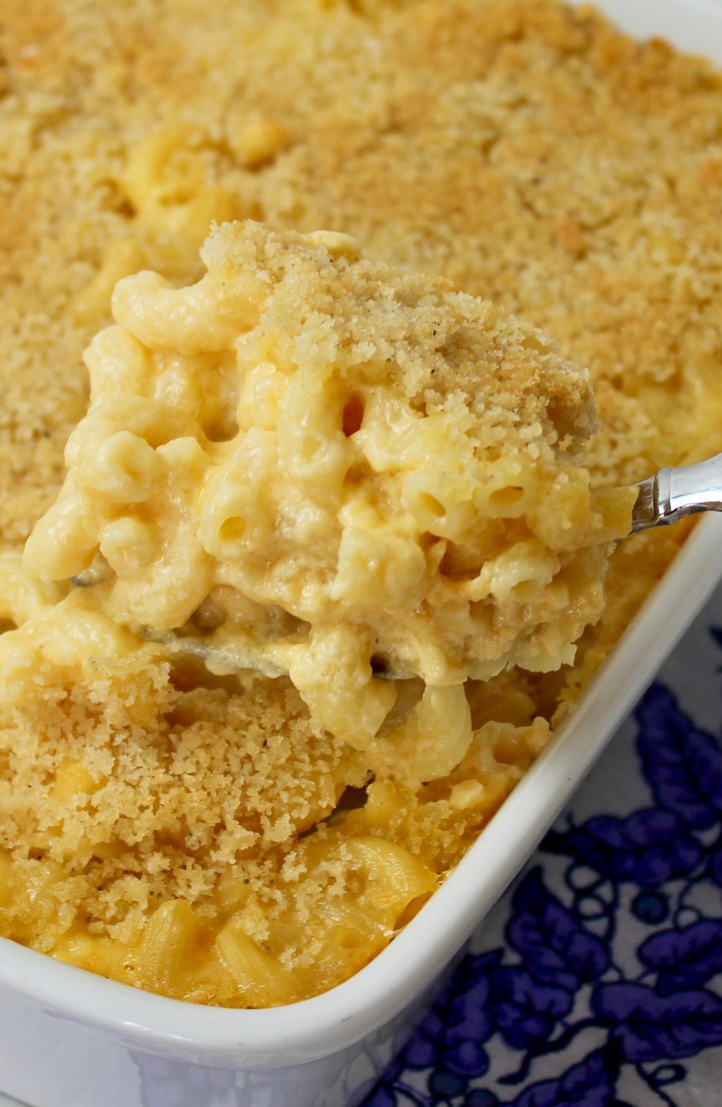 Recipe Photo - Portrait Orientation. Homemade Macaroni and Cheese in a white casserole stoneware dish. Set on a blue and white floral napkin. A silver serving spoon is scooping some of the creamy golden elbow macaroni. There is a golden brown panko crispy topping on top of the macaroni.