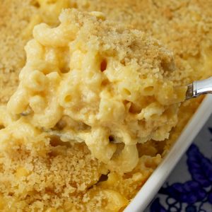 Recipe Photo - Portrait Orientation. Homemade Macaroni and Cheese in a white casserole stoneware dish. Set on a blue and white floral napkin. A silver serving spoon is scooping some of the creamy golden elbow macaroni. There is a golden brown panko crispy topping on top of the macaroni.