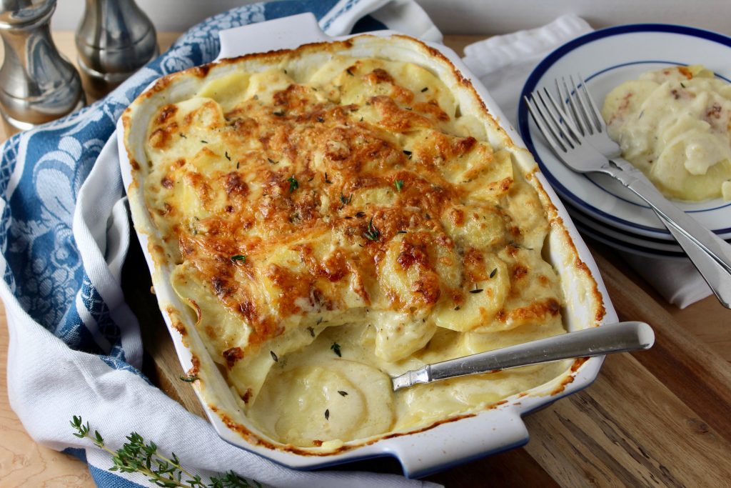Baked Casserole dish of Creamy Potatoes Au Gratin, 1 portion spooned out onto a plate. Blue and white patterned kitchen towel in the background.