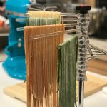 KitchenAid pasta drying rack with plain, tomato and spinach spaghetti and fettucini pasta hanging from rack. KitchenAid stand mixer with pasta roller attachment in background