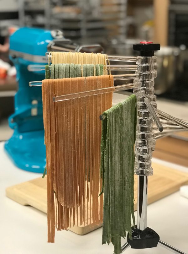KitchenAid pasta drying rack with plain, tomato and spinach spaghetti and fettucini pasta hanging from rack. KitchenAid stand mixer with pasta roller attachment in background