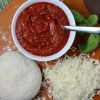 Bowl of easy homemade pizza sauce on pizza peel with shredded mozzarella, fresh pizza dough and sprig of fresh basil
