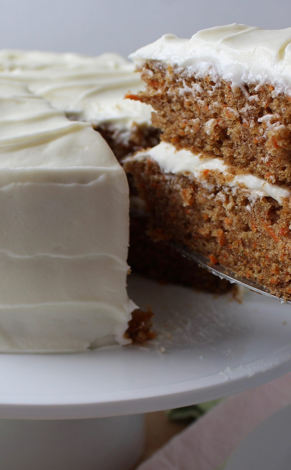 Recipe Photo for Cream Cheese Frosting Showing a 2 layer carrot cake frosted with cream cheese frosting on a white pedestal with a piece being lifted.