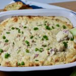 Chicken Cordon Bleu Dip in a rectangular blue casserole dish. Celery stick being scooped into hot dip sprinkled with sliced green onion