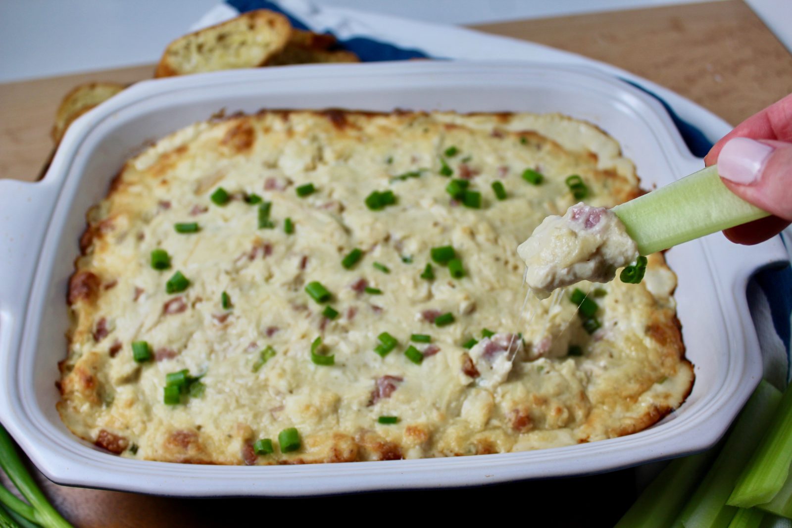 Chicken Cordon Bleu Dip in a rectangular blue casserole dish. Celery stick being scooped into hot dip sprinkled with sliced green onion