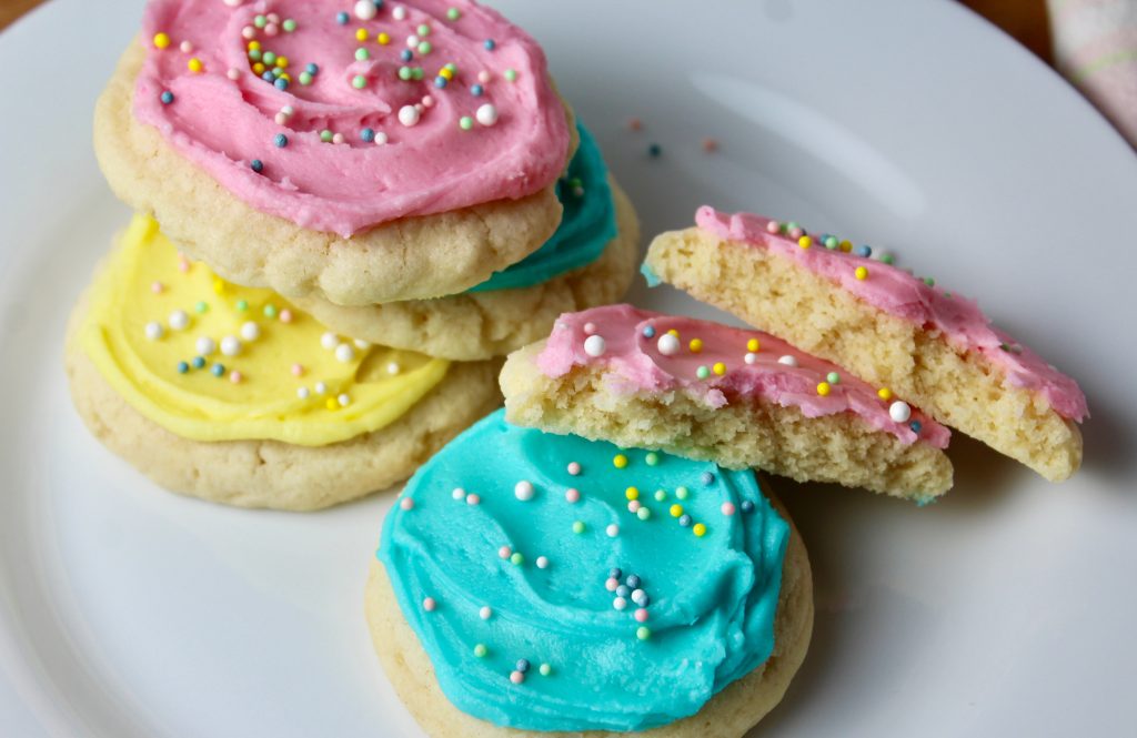 Cream Cheese Sugar cookies frosted in light pink, yellow and blue with nonpareil sprinkles - on white plate, one cookie broken in half
