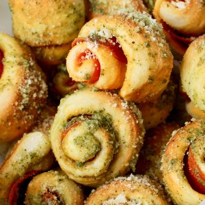Recipe Photo for Pizzeria Style Pepperoni Bites. Basket with waxed paper liner filled with rolls made of pizza dough, filled with pepperoni, baked and drizzle with fresh basil pesto and Parmesan Cheese.