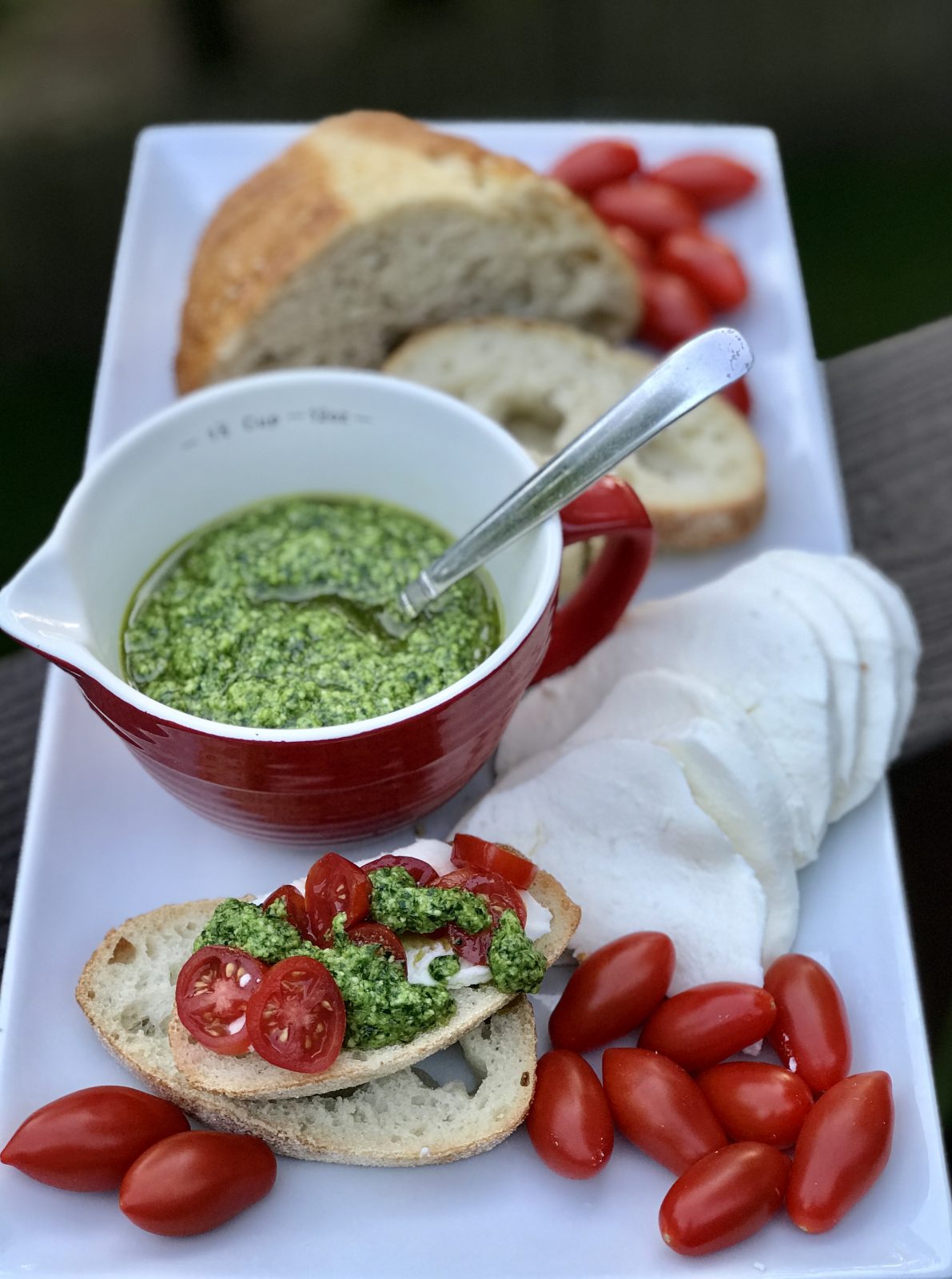 White rectangular tray with bread, spinach basil pesto in a red cup, mozzarella and grape tomatoes
