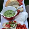 White rectangular tray with bread, spinach basil pesto in a red cup, mozzarella and grape tomatoes