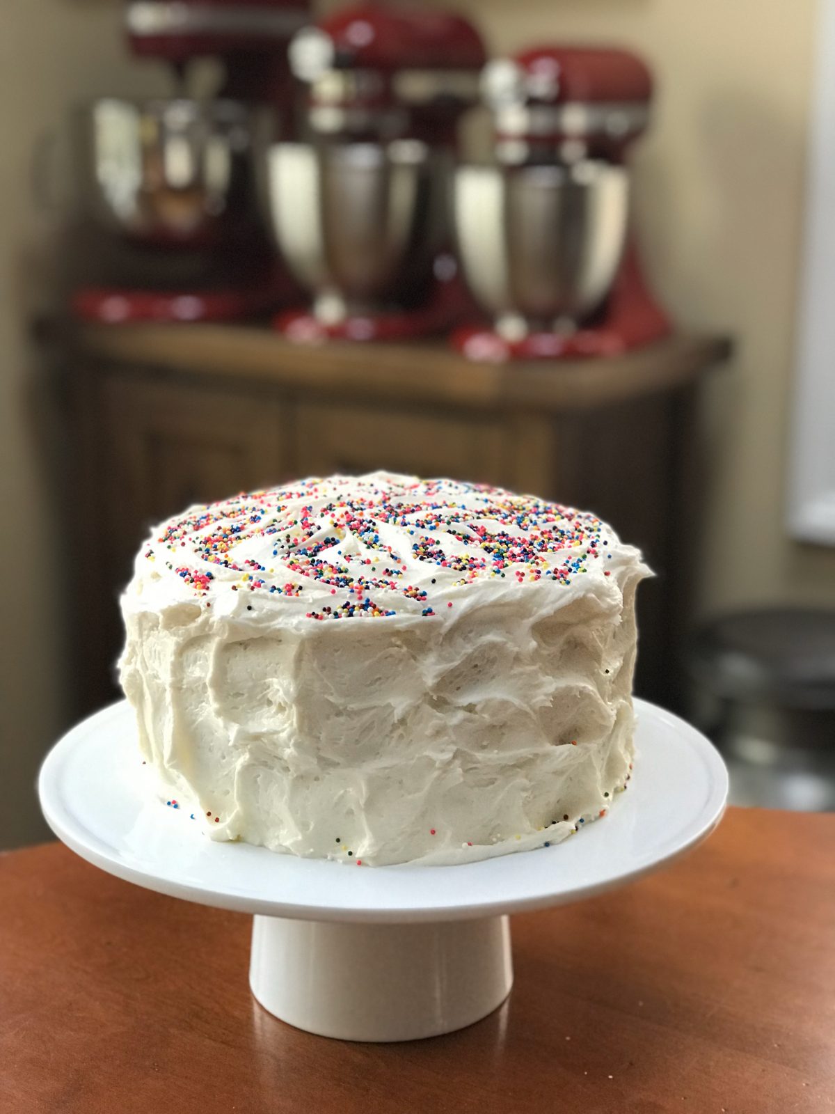 Frosted yellow cake on pedestal with sprinkles and KitchenAid Mixers in background