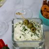 Recipe Photo for Dirty Martini Dip. Shows the white creamy dip sprinkled with fresh snipped chives and a bamboo pick with 3 olives. In the background are ramekins with pretzel chips and carrots and cucumber slices. Also 2 martini glasses with dirty martinis.