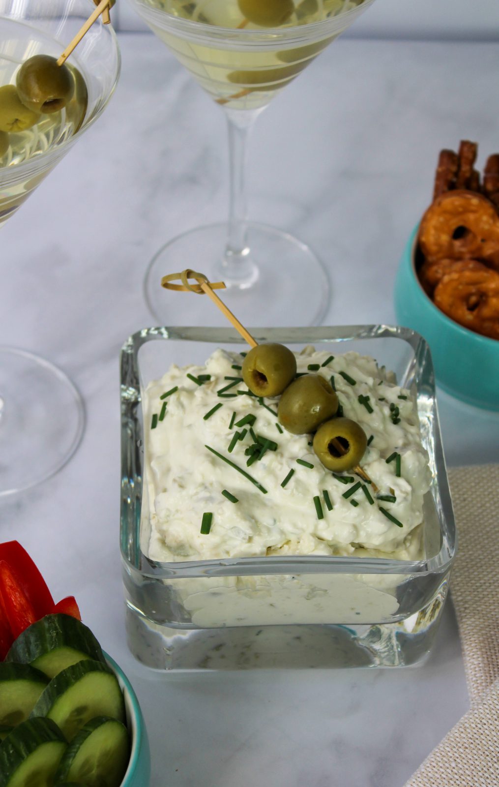 Recipe Photo for Dirty Martini Dip. Shows the white creamy dip sprinkled with fresh snipped chives and a bamboo pick with 3 olives. In the background are ramekins with pretzel chips and carrots and cucumber slices. Also 2 martini glasses with dirty martinis.