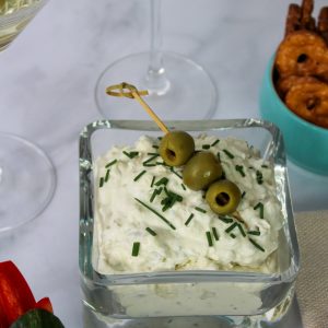 Recipe Photo for Dirty Martini Dip. Shows the white creamy dip sprinkled with fresh snipped chives and a bamboo pick with 3 olives. In the background are ramekins with pretzel chips and carrots and cucumber slices. Also 2 martini glasses with dirty martinis.