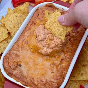 Small rectangular dish with cheese topped hot queso dip on wooden platter with tortilla chips and red pepper strips