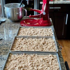Vertical oriented photo with red 7 quart KitchenAid Stand Mixer in background and 3, 13 x 9 inch metal baking pans with coffee cake and brown sugar crumble ready to be baked.