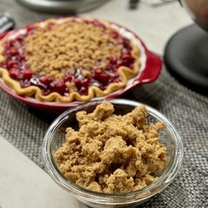 Vertical photo featuring brown sugar crumble in a bowl in the foreground, a cherry pie with brown sugar crumble in the middle of the photo and a KitchenAid Stand Mixer faded out in the background.