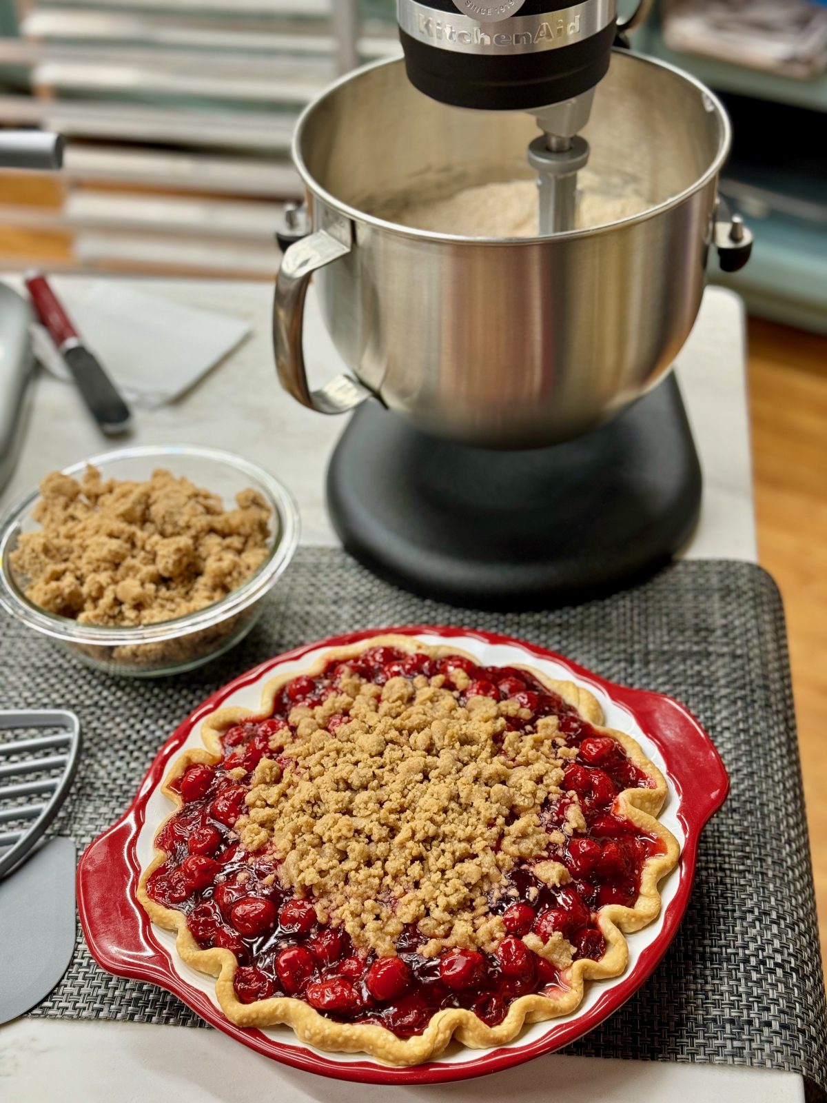 Vertical photo with a cherry pie with brown sugar crumble topping in the foreground. A bowl of brown sugar crumble topping in the middle of the photo and a KitchenAid stand mixer in the background.
