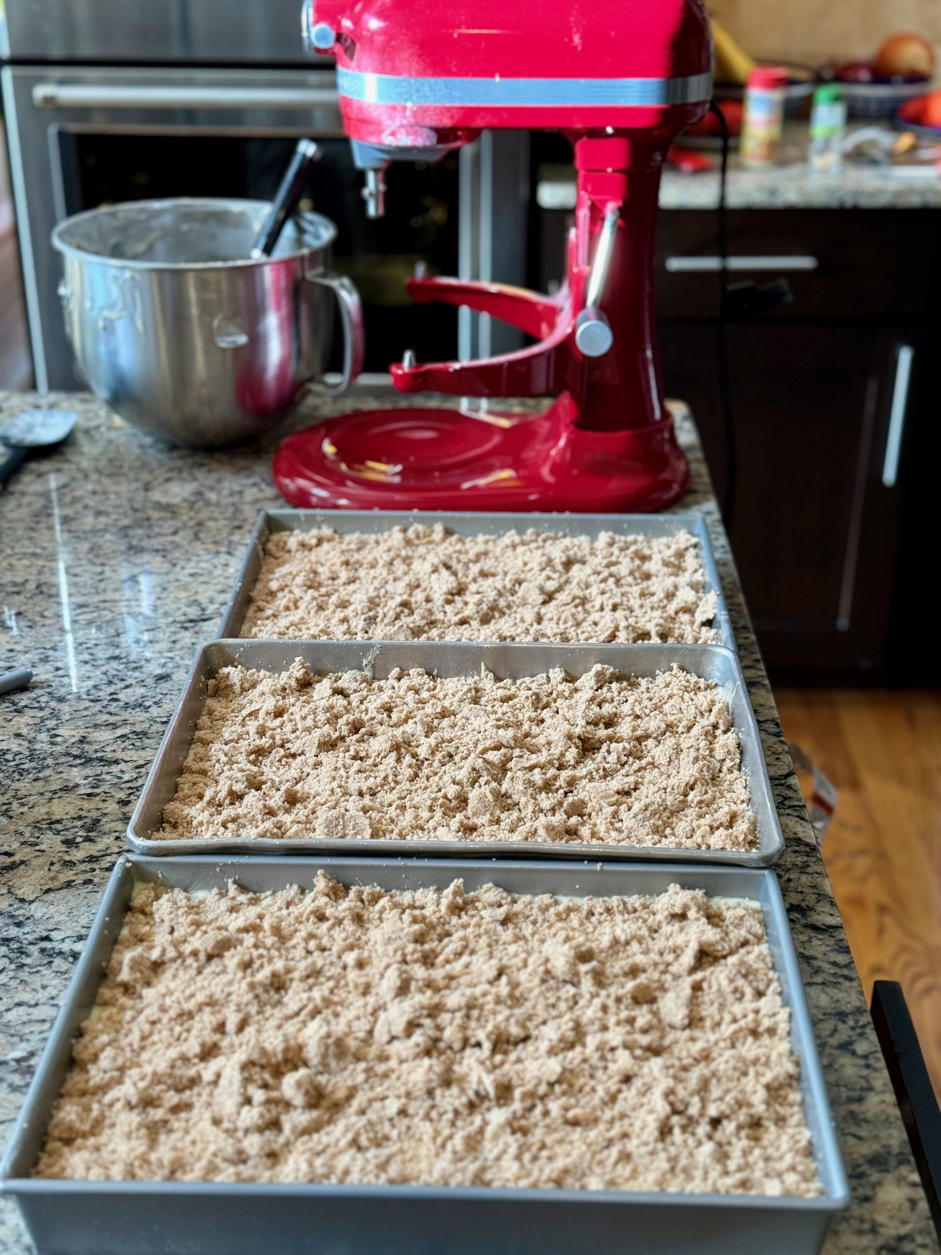 Vertical oriented photo with red 7 quart KitchenAid Stand Mixer in background and 3, 13 x 9 inch metal baking pans with coffee cake and brown sugar crumble ready to be baked.