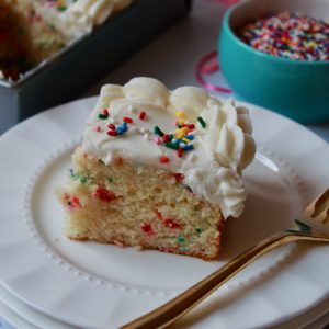 One corner slice of white frosted sheet cake with colorful sprinkles on a stack of three plates with a gold fork. Sheet cake and bowls of sprinkles in the background.