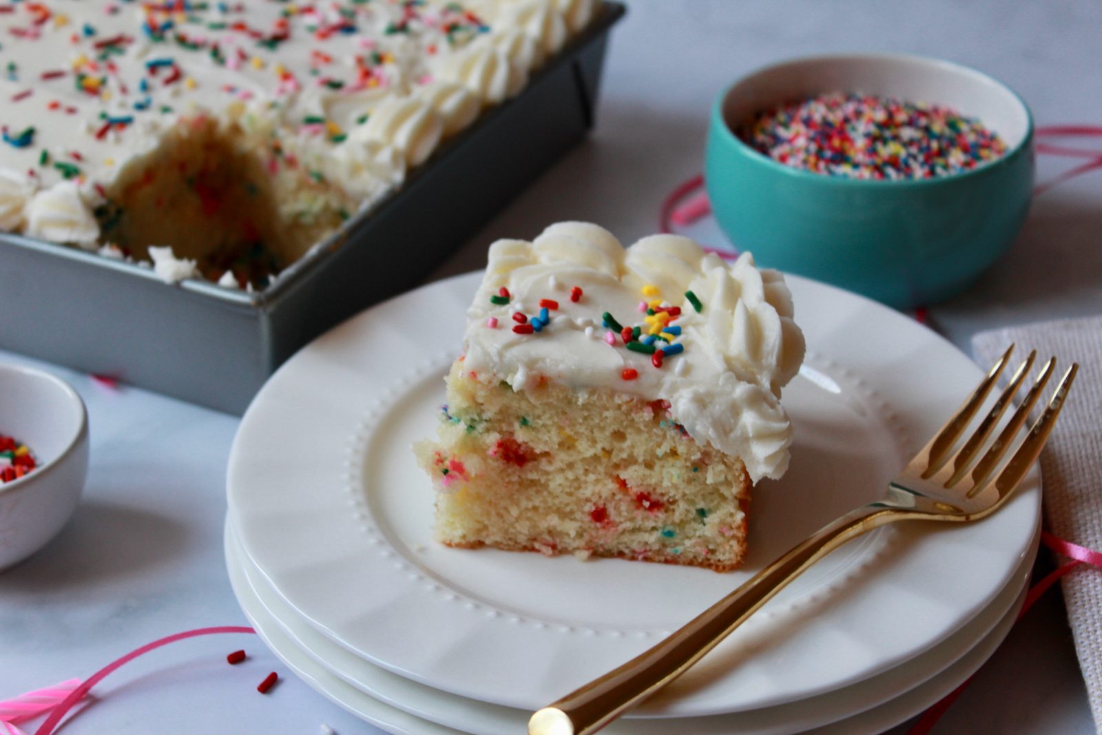 A horizontal oriented photo of a sprinkle vanilla sheet cake. The metal pan containing the cake with a slice cut out is in the background with a slice of cake in the foreground on a stack of white plates with a gold fork. On either side there are small bowls of sprinkles with some spilled on the white counter surface and a length of pink ribbon.