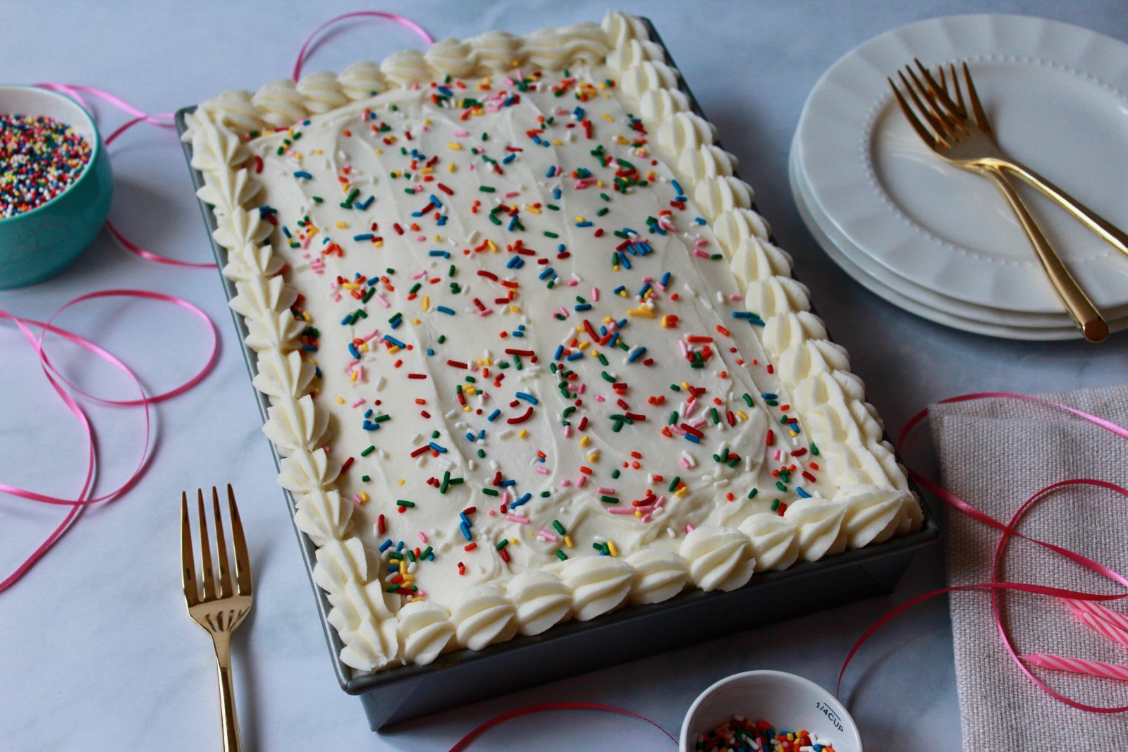 A horizontal oriented photo - looking down upon a sprinkle vanilla sheet cake.  The cake is in a rectangular metal pan and is uncut.  The cake is frosted in vanilla frosting with a piped border and topped with colorful sprinkles.  Around the cake are pink birthday candles, gold forks, white dessert plates, small bowls of sprinkles as well as thin pink ribbon and some strewn sprinkles.