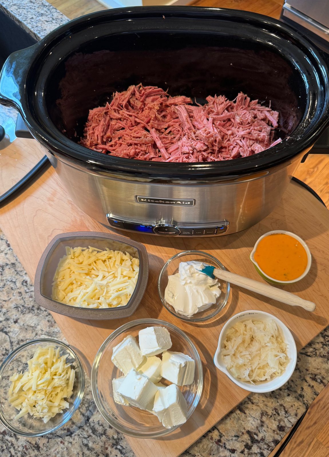 All the ingredients to make Slow Cooker Reuben Dip.  In the back is a KitchenAid slow cooker with shredded corned beef.  In front on a wooden board are small dishes of shredded swiss cheese, cream cheese chunks, sour cream and Russian Dressing
