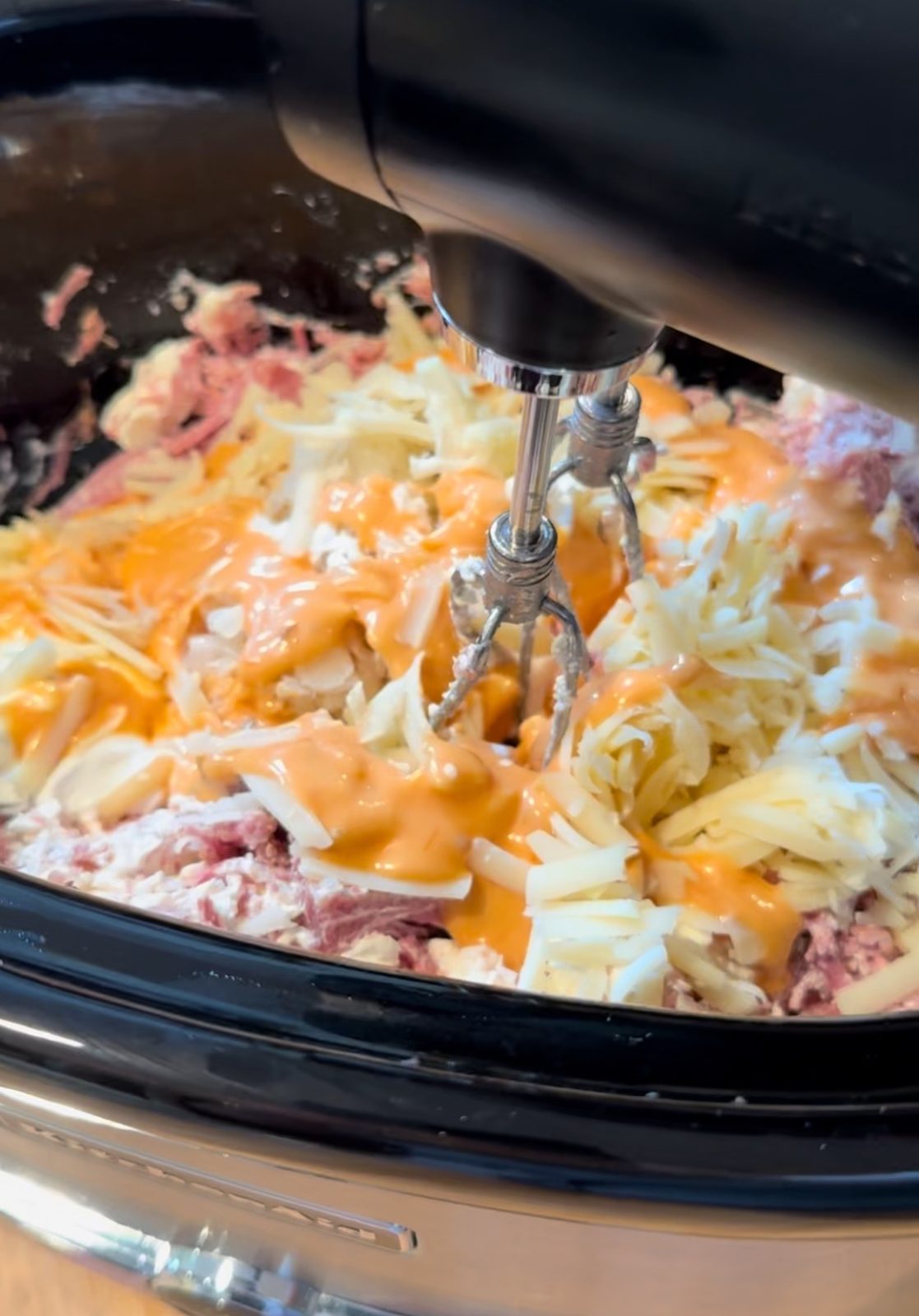 Making Slow Cooker Reuben Dip.  Close up photo of black slow cooker with shredded corned beef, sauerkraut, cream cheese, sour cream, Russian Dressing and Swiss cheese being mixed with a black KitchenAid Cordless hand mixer
