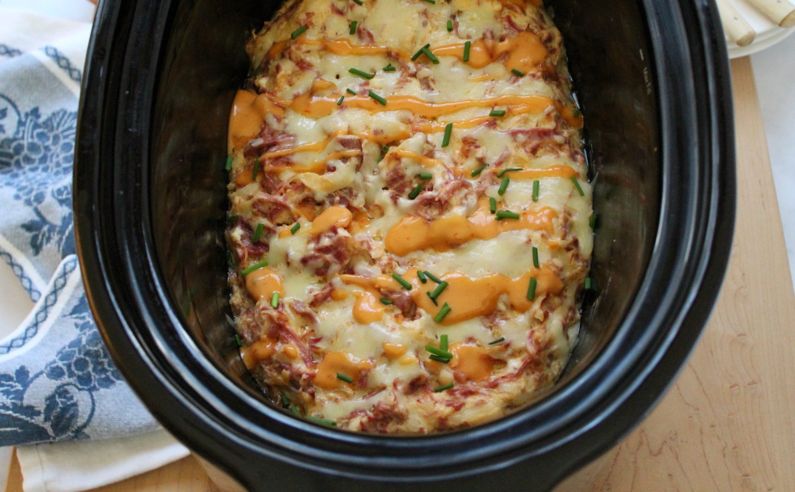 Close up shot of Reuben Dip in a black slow cooker.  The dip is topped with extra melted swiss cheese, a drizzle of Russian dressing and a sprinkling of snipped chives.