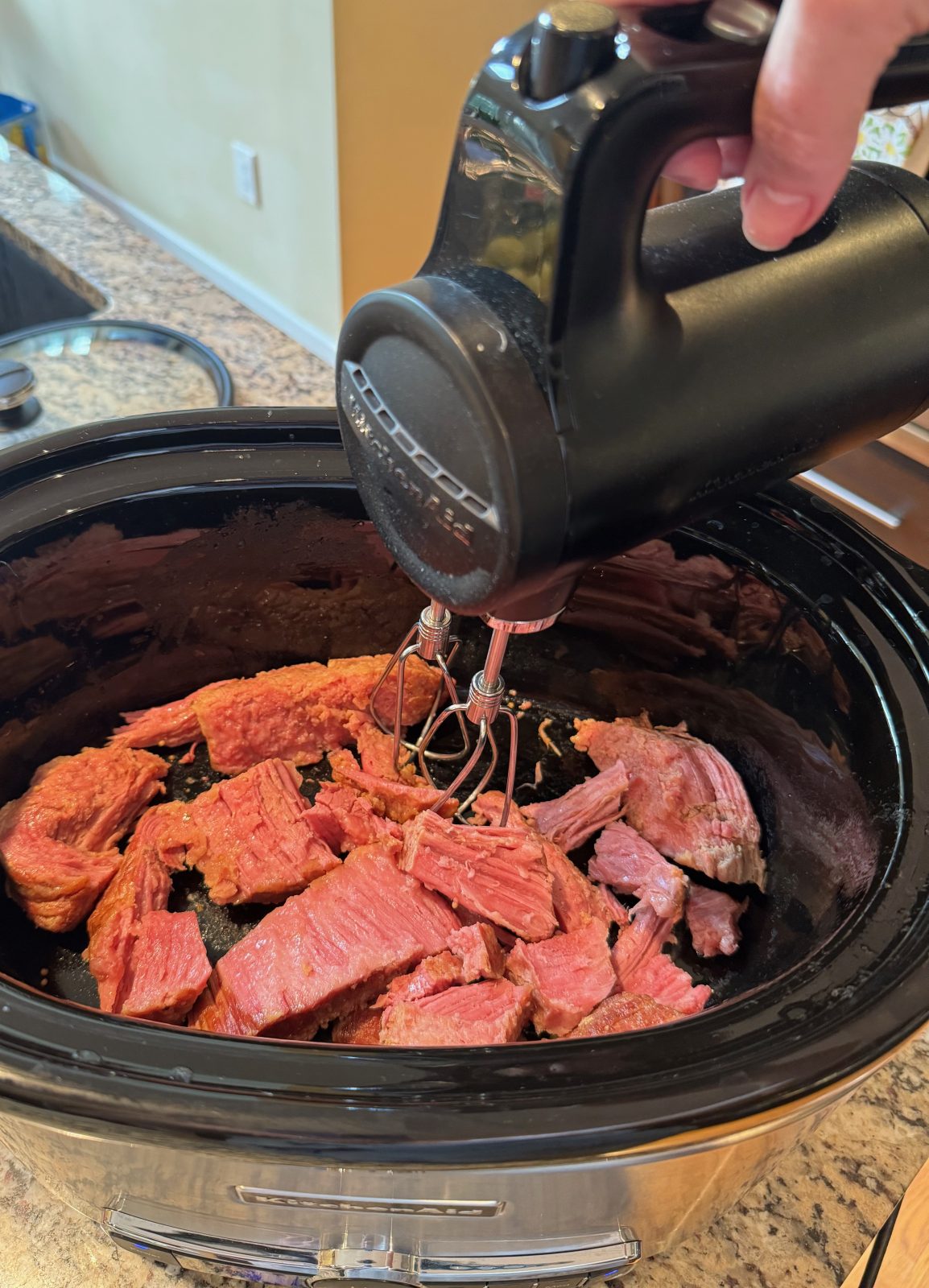 Starting to make Slow Cooker Reuben Dip.  I have 1 pound of cooked corned beef pieces in a black slow cooker with black cordless KitchenAid hand mixer getting ready to shred the corned beef.
