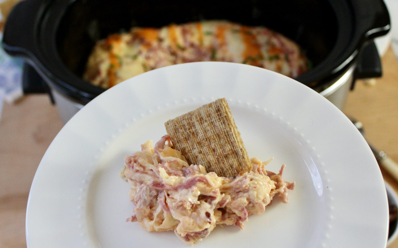 A white plate with a dollop of Slow Cooker Reuben Dip and a Triscuit cracker stuck into the dollop.  In the background is the black slow cooker containing the Slow Cooker Reuben Dip.
