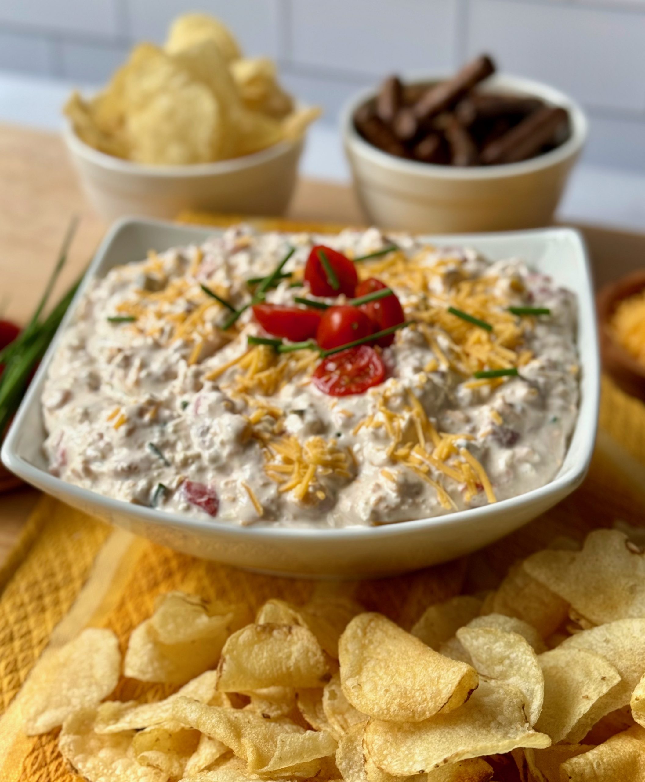 Vertical recipe photo of Sink the Boat Dip. Sloped white rectangular bowl in center with dip, creamy white dip topped with shredded yellow cheddar, halved grape tomatoes and snipped chives. Surrounding the dip are potato chips and pumpernickel sticks. The backsplash is white subway tile.