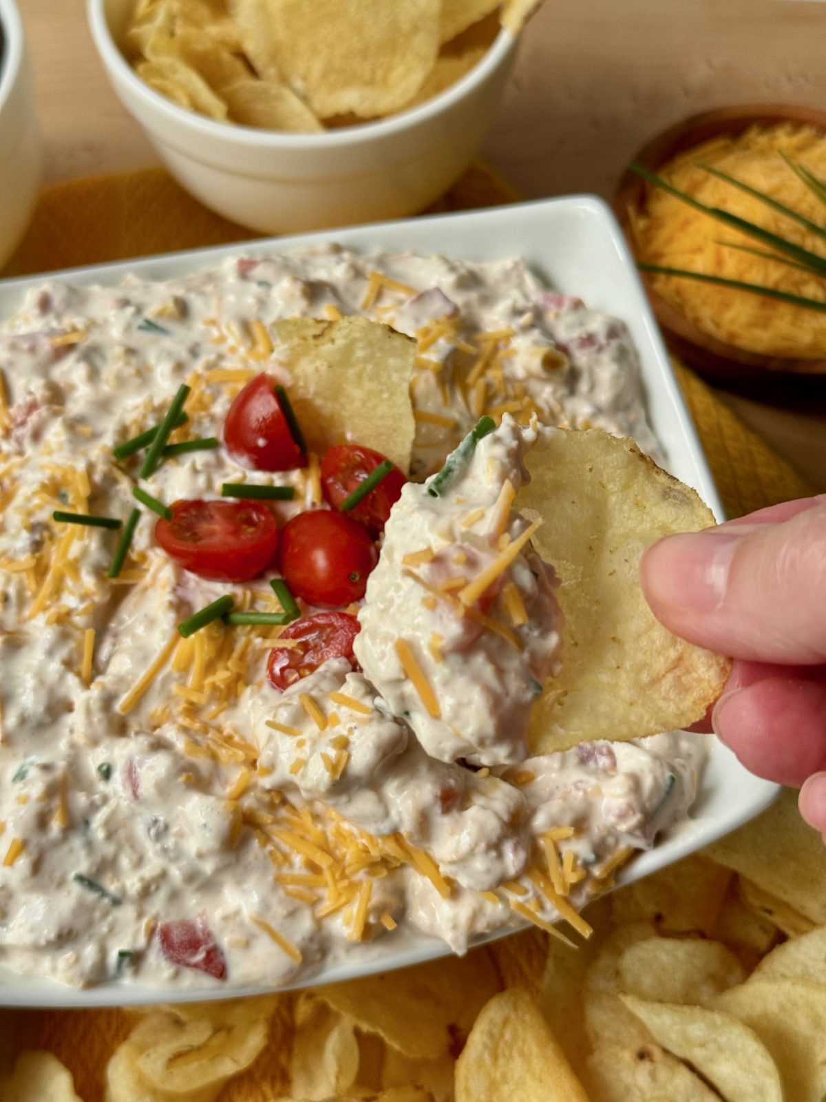 Vertical recipe photo of Sink the Boat Dip. Close up of white rectangular bowl filled with creamy colored dip topped with halved grape tomatoes and chives. A hand is holding a scoop of dip on a potato chip. In the background are some potato chips and a small wooden dish with shredded cheddar and chives.