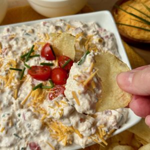 Vertical recipe photo of Sink the Boat Dip. Close up of white rectangular bowl filled with creamy colored dip topped with halved grape tomatoes and chives. A hand is holding a scoop of dip on a potato chip. In the background are some potato chips and a small wooden dish with shredded cheddar and chives.