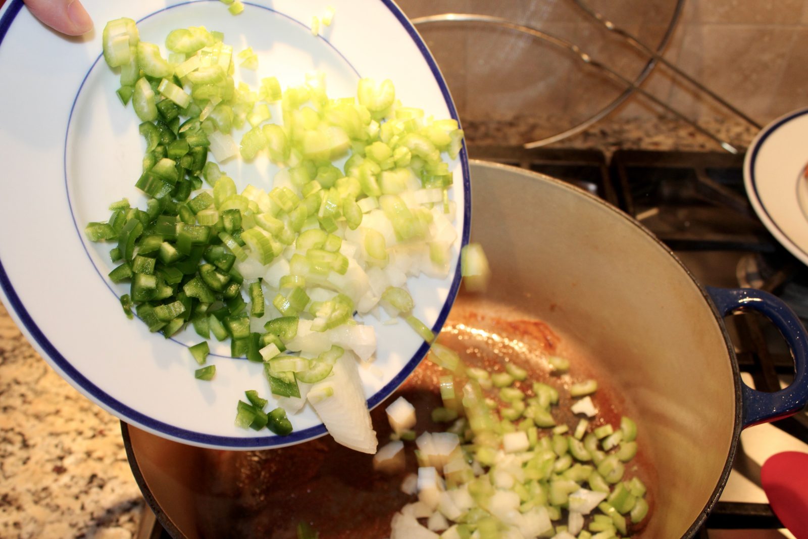 Recipe photo for White Chicken Chili showing chopped celery, onion and jalapeño being slid into pot from white plate with blue ring. The pot is on a gas stove.