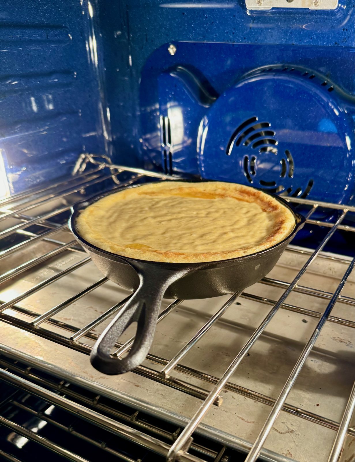 recipe photo for sourdough cornbread showing cornbread batter baking in oven with blue interior. the edges of the cornbread are puffed up inside the 8-inch cast iron skillet