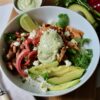 Overhead image of chicken salsa rice bowl with sliced avocado, pinto beans, cilantro, pickled red onions, queso fresco and a dollop of avocado ranch sauce. The dish is in a white round ceramic bowl on top of a wooden board. There is a dish of additional sauce, cilantro and avocado in the background.