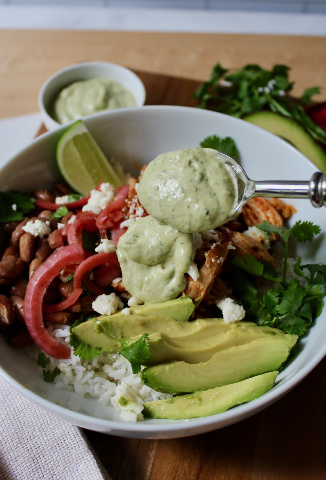 Overhead image of chicken salsa rice bowl with sliced avocado, pinto beans, cilantro, pickled red onions, queso fresco and a spoon with a dollop of avocado ranch sauce being added to the top.  The dish is in a white round ceramic bowl on top of a wooden board.  There is a dish of additional sauce, cilantro and avocado in the background.
