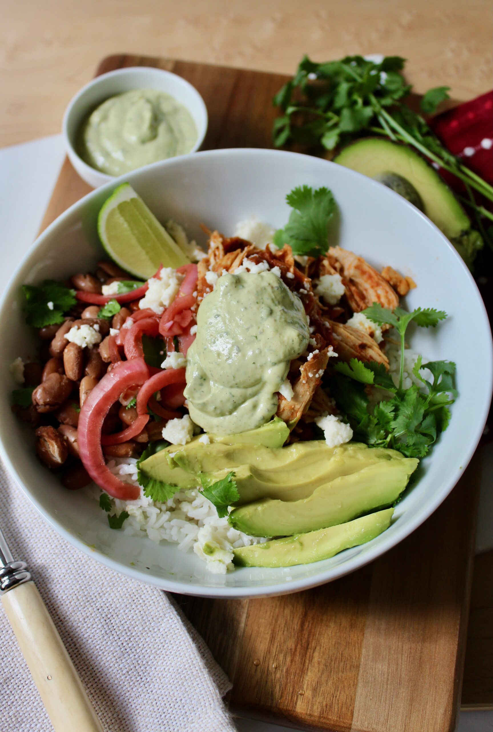 Overhead image of chicken salsa rice bowl with sliced avocado, pinto beans, cilantro, pickled red onions, queso fresco and a dollop of avocado ranch sauce. The dish is in a white round ceramic bowl on top of a wooden board. There is a dish of additional sauce, cilantro and avocado in the background.