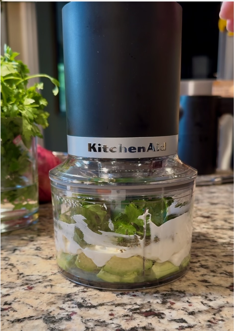 image of front of KitchenAid Top Down Cordless Chopper in black matte.  The bowl is filled with the ingredients to make Avocado Ranch Sauce.  There is a granite countertop, vase of cilantro and oven in the background.